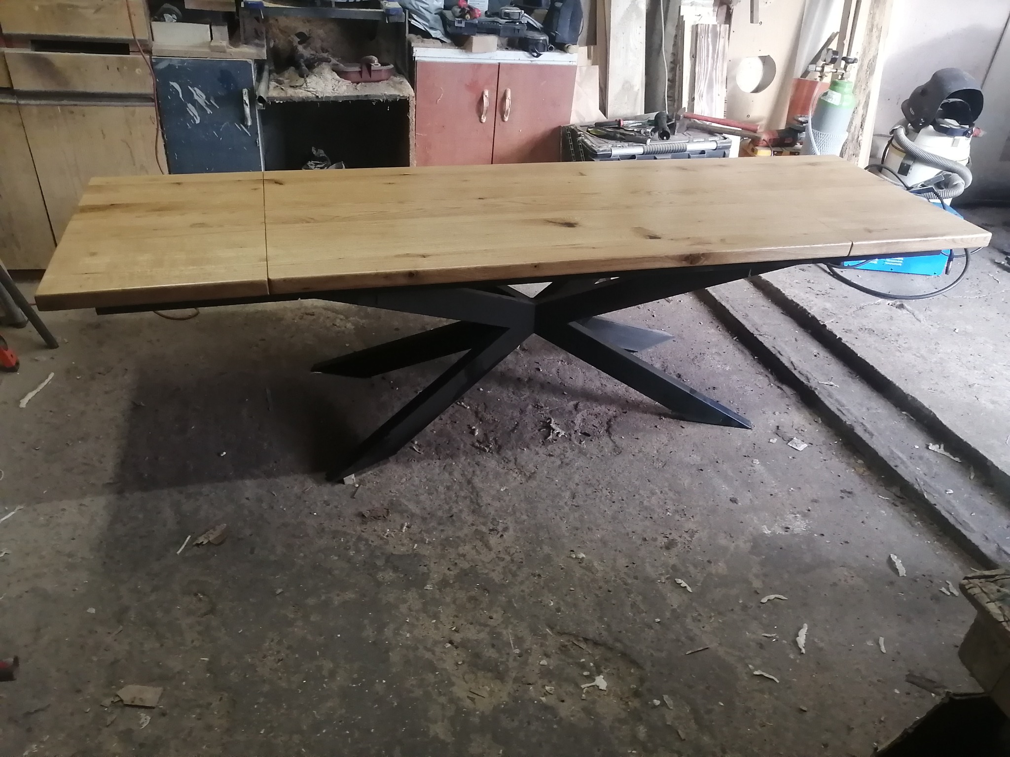 Rozkładany stół z blatem z jasnego drewna i czarną, geometryczną podstawą. Stół stoi w warsztacie, widoczne narzędzia i materiały stolarskie w tle.