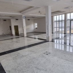 Voitech Studio - Przestronne wnętrze w trakcie wykańczania: jasnoszara, wzorzysta podłoga z ciemnymi obramowaniami, kolumny z widocznymi śladami prac, sufit z wbudowanymi klimatyzatorami i wiszącymi przewodami.