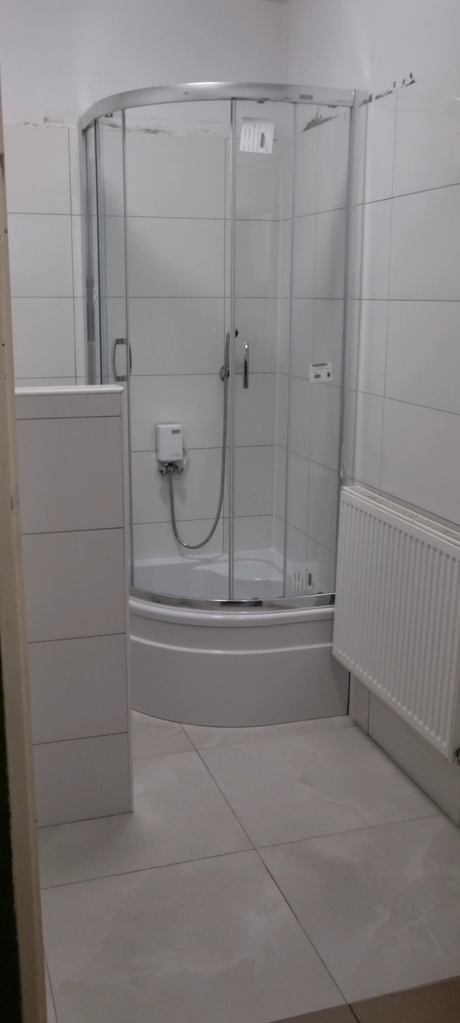 Narożna kabina prysznicowa z brodzikiem, białe płytki na ścianach i podłodze, grzejnik na ścianie, widoczne ślady niedokończonych prac wykończeniowych na łączeniu płytek i kabiny.