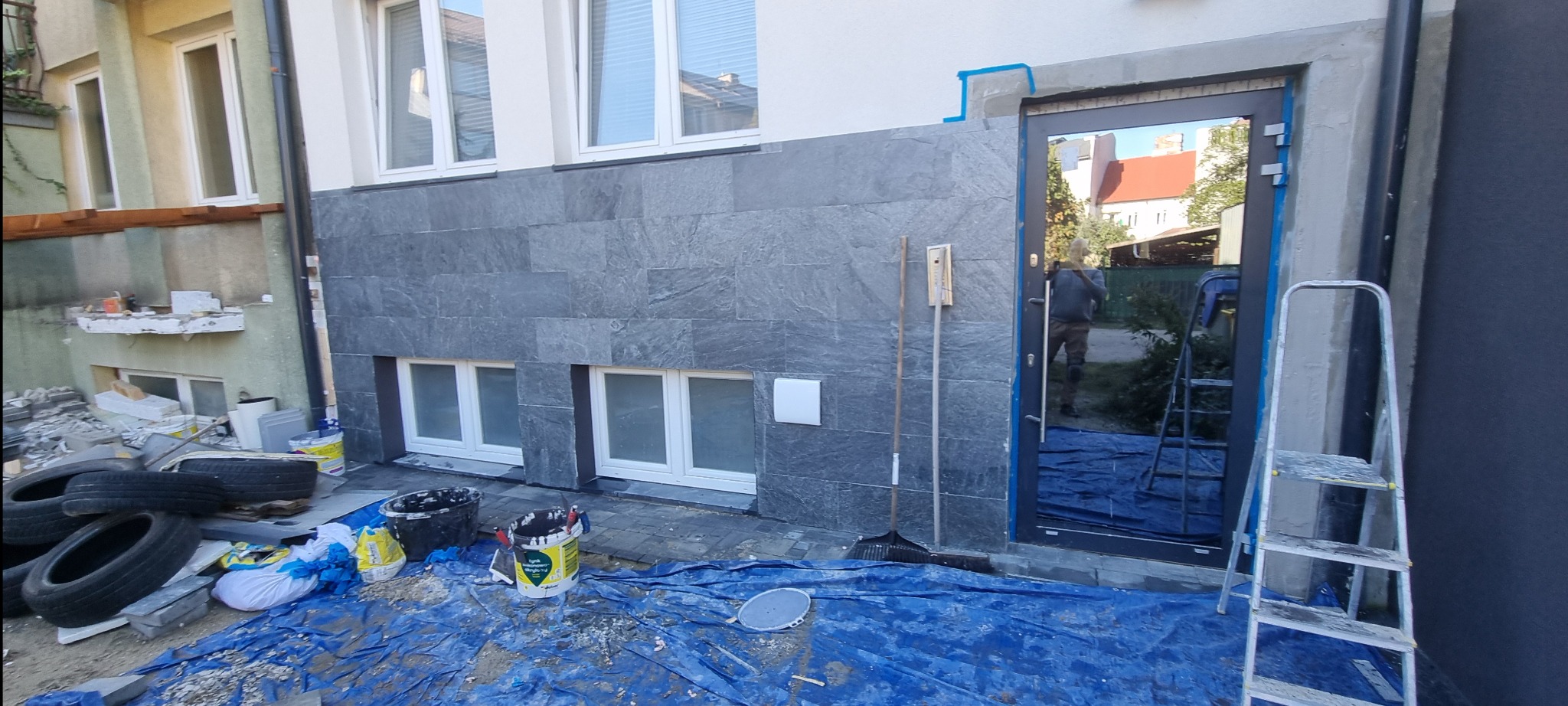 Elewacja budynku z szarej, kamiennej okładziny w trakcie montażu, widoczne narzędzia, wiadro z farbą i folia ochronna na ziemi.