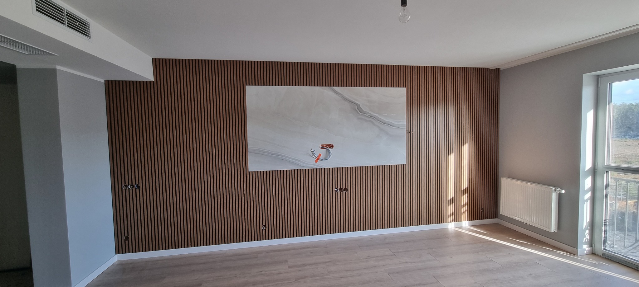 Pusty pokój z drewnianą ścianą z listew, na której wisi płyta imitująca marmur z wystającymi kablami elektrycznymi, szare ściany boczne i jasna podłoga.