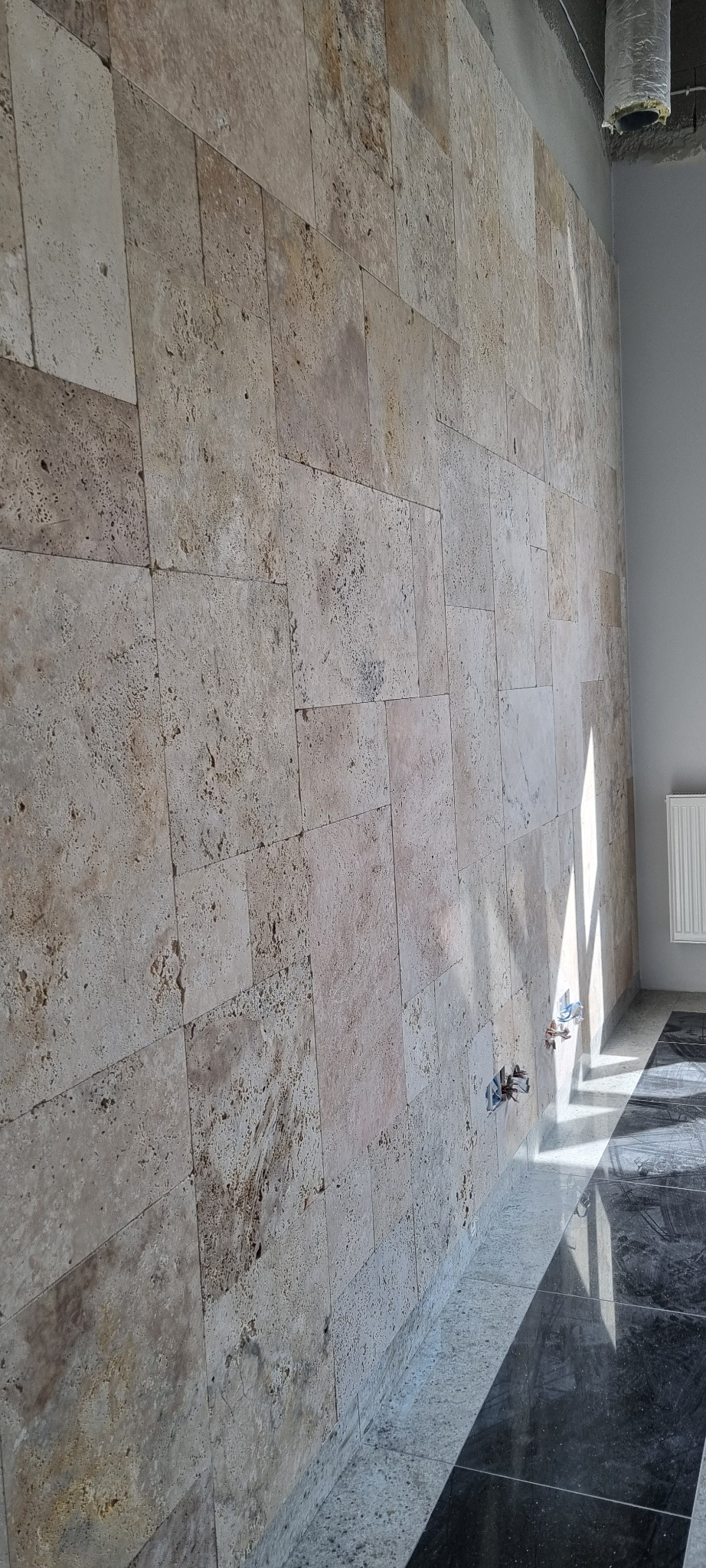 Ściana wyłożona beżowymi, porowatymi płytkami trawertynowymi, z widocznymi otworami na instalacje elektryczne, na tle błyszczącej, czarno-białej podłogi.