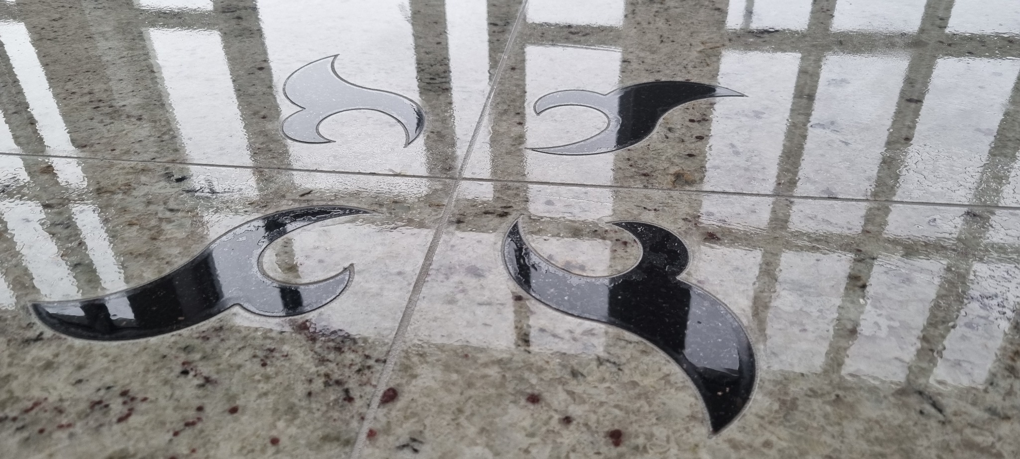 Polerowana, granitowa podłoga z wkomponowanymi, czarnymi, abstrakcyjnymi wzorami w kształcie płomieni lub łez, odbijająca światło i krople wody.