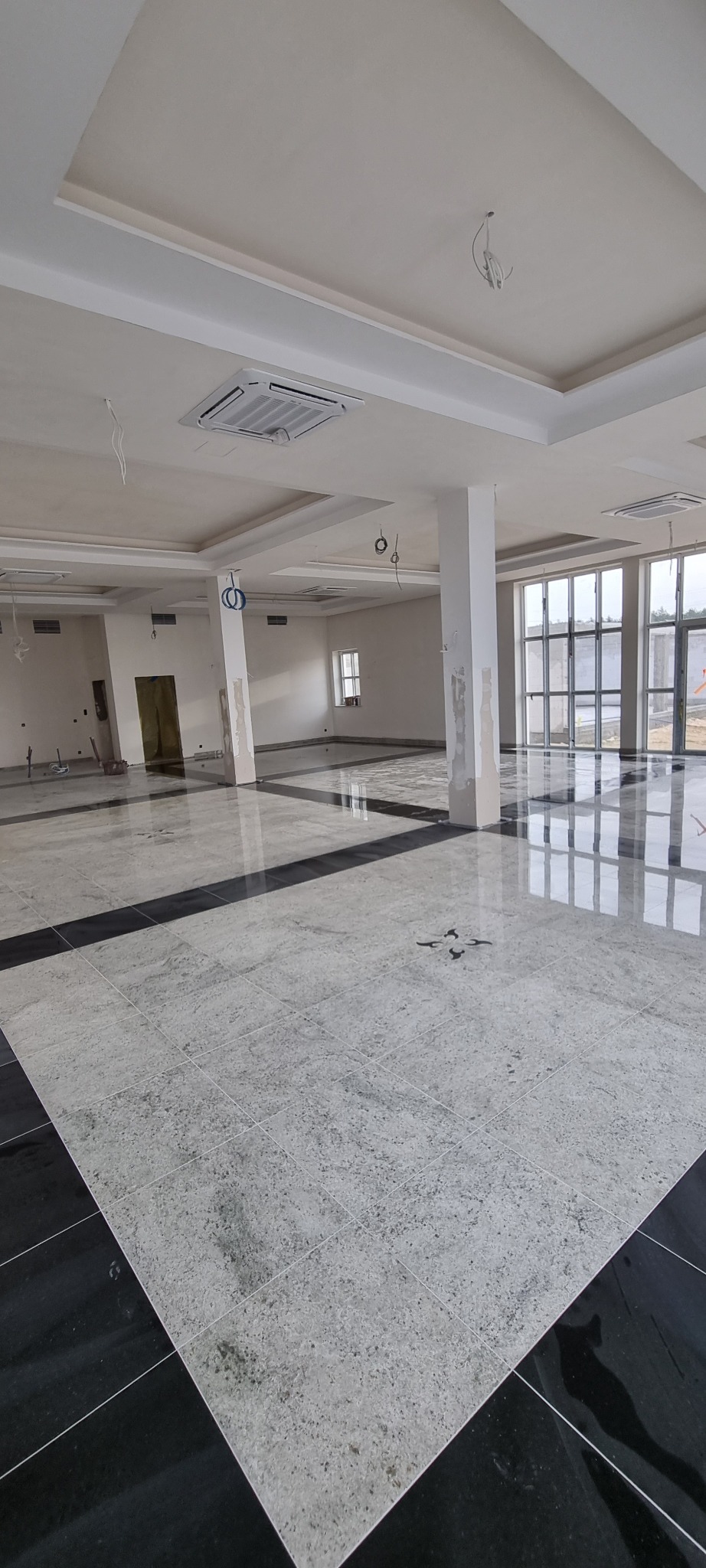 Przestronne wnętrze w trakcie wykańczania: jasnoszara, wzorzysta podłoga z ciemnymi obramowaniami, kolumny z widocznymi śladami prac, sufit z wbudowanymi klimatyzatorami i wiszącymi przewodami.