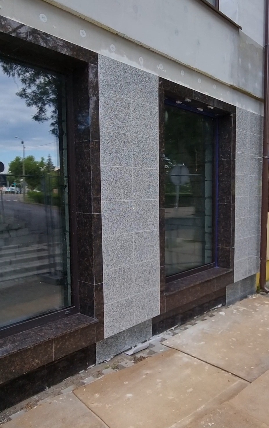 Elewacja budynku z oknami obłożona płytkami imitującymi kamień w dwóch odcieniach szarości, widoczne ślady po pracach wykończeniowych na chodniku.