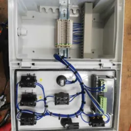 Otwarta skrzynka elektryczna z zamontowanymi elementami, widoczne złącza, przewody niebieskie i białe oraz listwy zaciskowe, narzędzia obok.