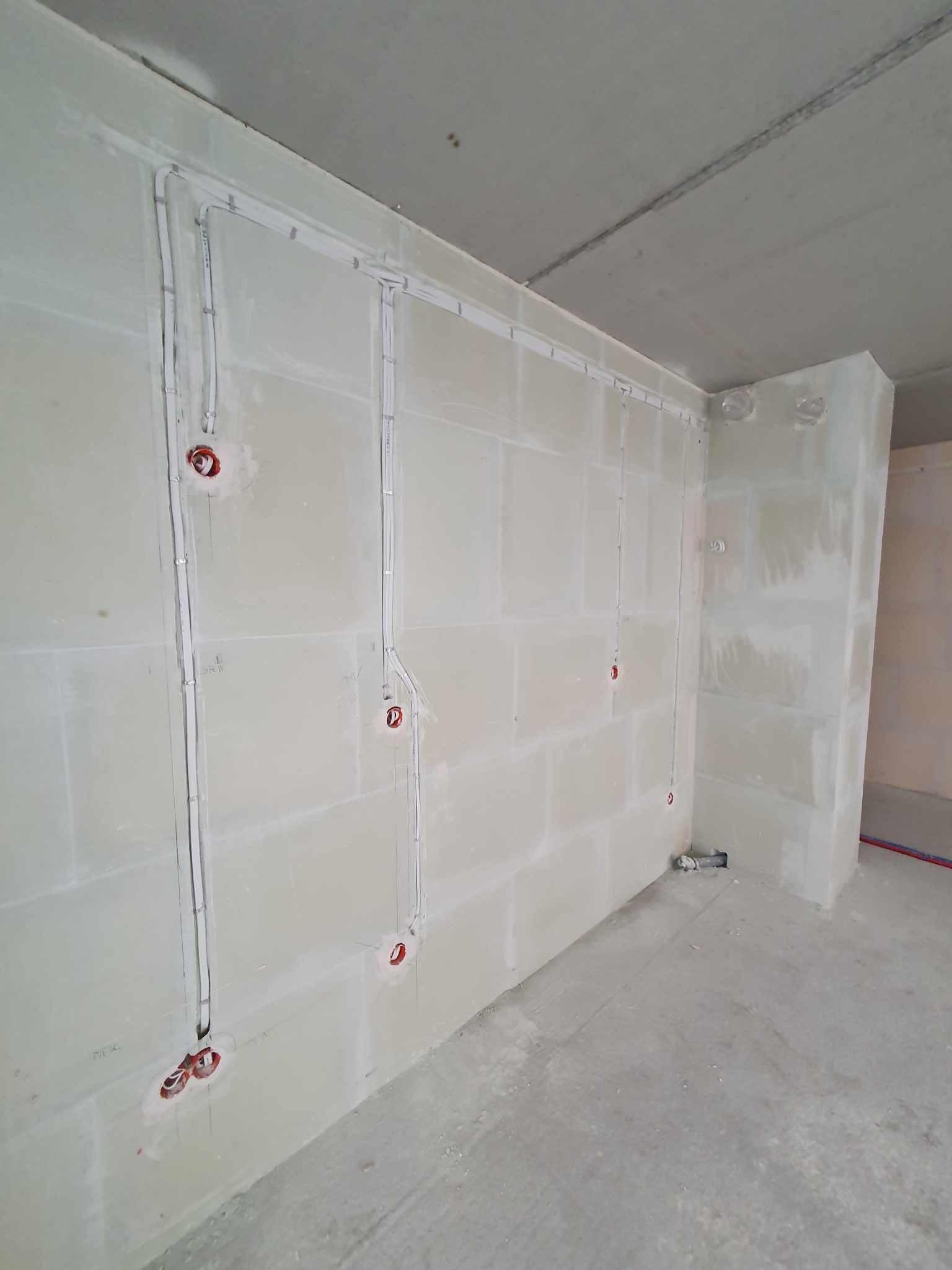 Wewnętrzne ściany w surowym stanie z zamontowanymi puszkami elektrycznymi i przewodami w białych listwach, widoczne także rury instalacji wodnej przy podłodze.