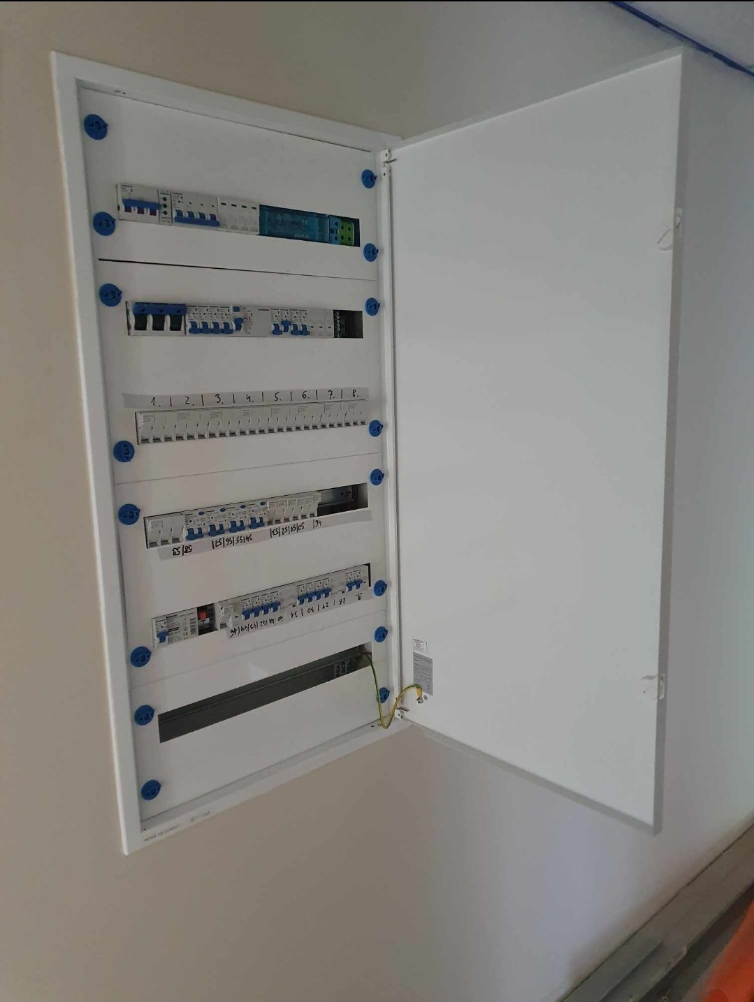 Otwarta skrzynka elektryczna z bezpiecznikami i przewodami, zamontowana na ścianie wewnątrz budynku.