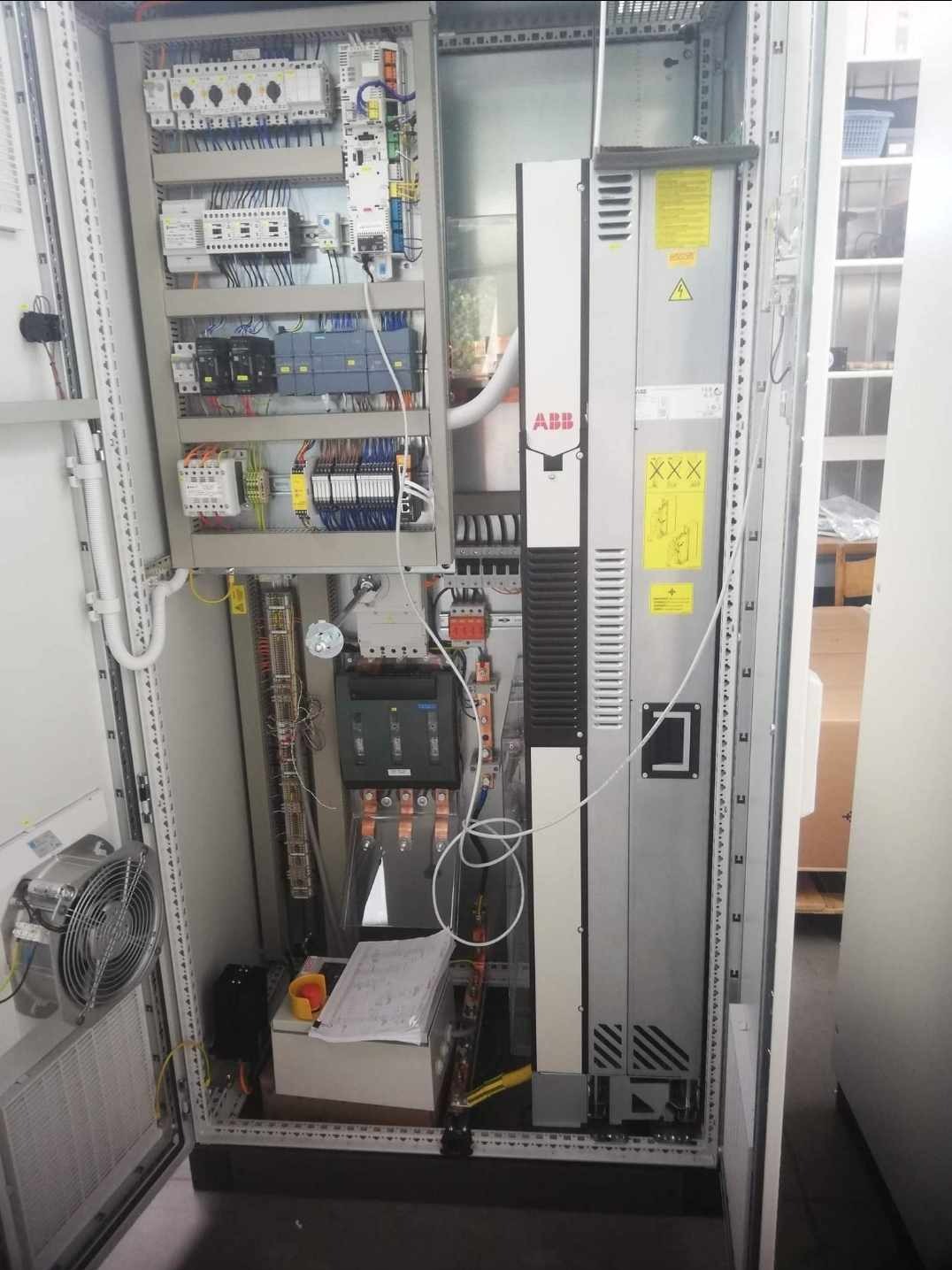 Otwarta szafa sterownicza z widocznymi elementami elektrycznymi, w tym modułem ABB, zabezpieczeniami, okablowaniem i wentylatorem na drzwiach; schemat elektryczny na białej skrzynce w dolnej części.