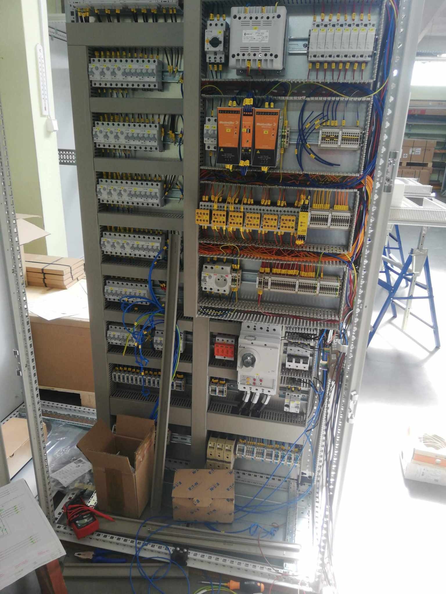 Szafa sterownicza w trakcie montażu, widoczne liczne przewody, moduły, bezpieczniki i schemat elektryczny na podłodze obok narzędzi.