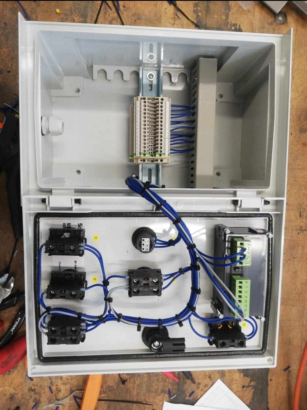 Otwarta skrzynka elektryczna z zamontowanymi elementami, widoczne złącza, przewody niebieskie i białe oraz listwy zaciskowe, narzędzia obok.
