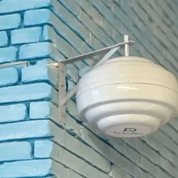 Biały, kulisty osuszacz powietrza marki RenoDry zamontowany na metalowym wsporniku na rogu błękitnej, pomalowanej ściany z cegieł.