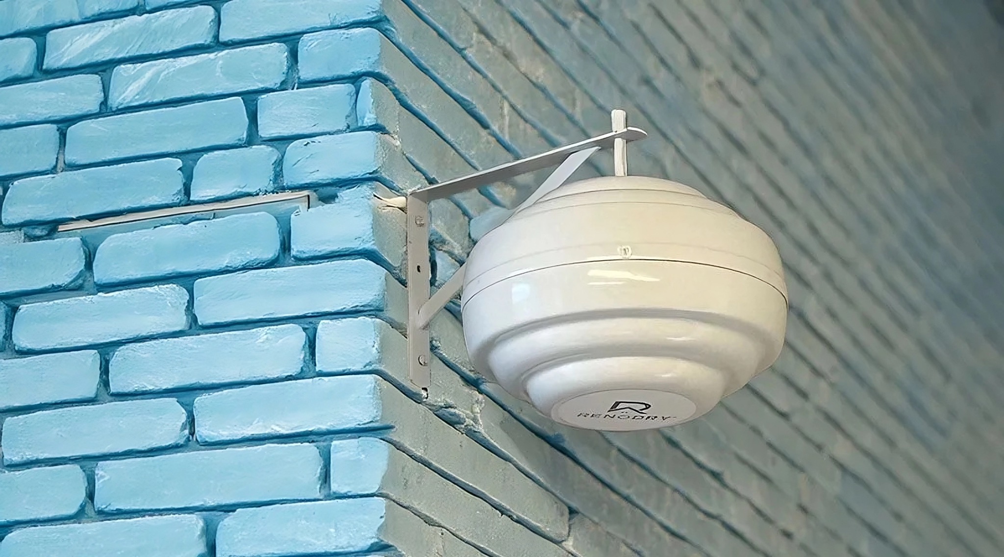 Biały, kulisty osuszacz powietrza marki RenoDry zamontowany na metalowym wsporniku na rogu błękitnej, pomalowanej ściany z cegieł.