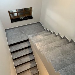 R.W. KONTRA Maciej Sowa - Schody wewnętrzne wykończone szarymi płytkami imitującymi kamień, z kontrastującymi czarnymi wstawkami na stopniach, oświetlone od dołu, z małym okienkiem w tle.