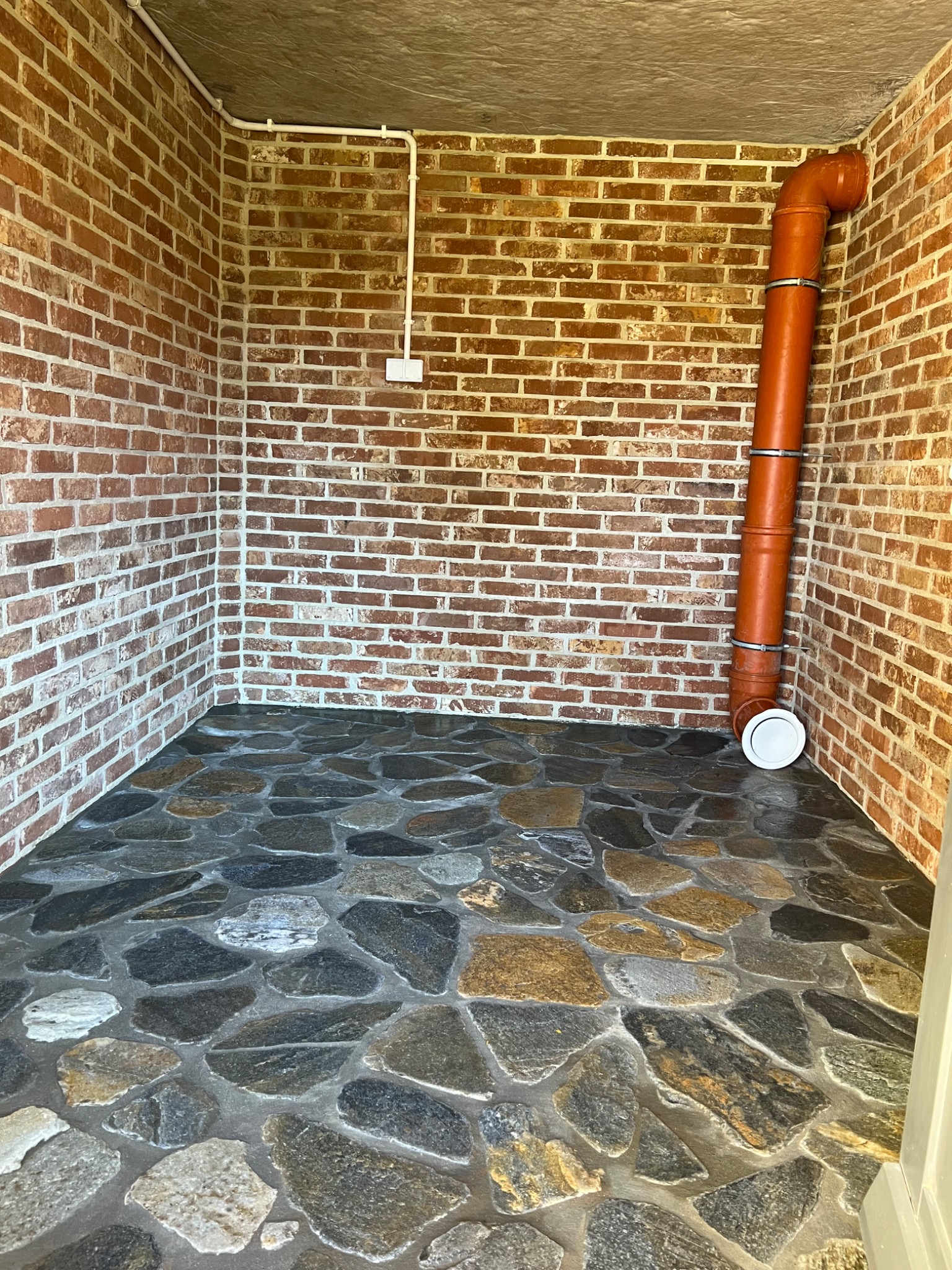 Wnętrze pomieszczenia z ceglanymi ścianami, podłogą wyłożoną nieregularnymi kamieniami w odcieniach szarości i brązu, oraz pomarańczową rurą kanalizacyjną biegnącą wzdłuż jednej ze ścian.