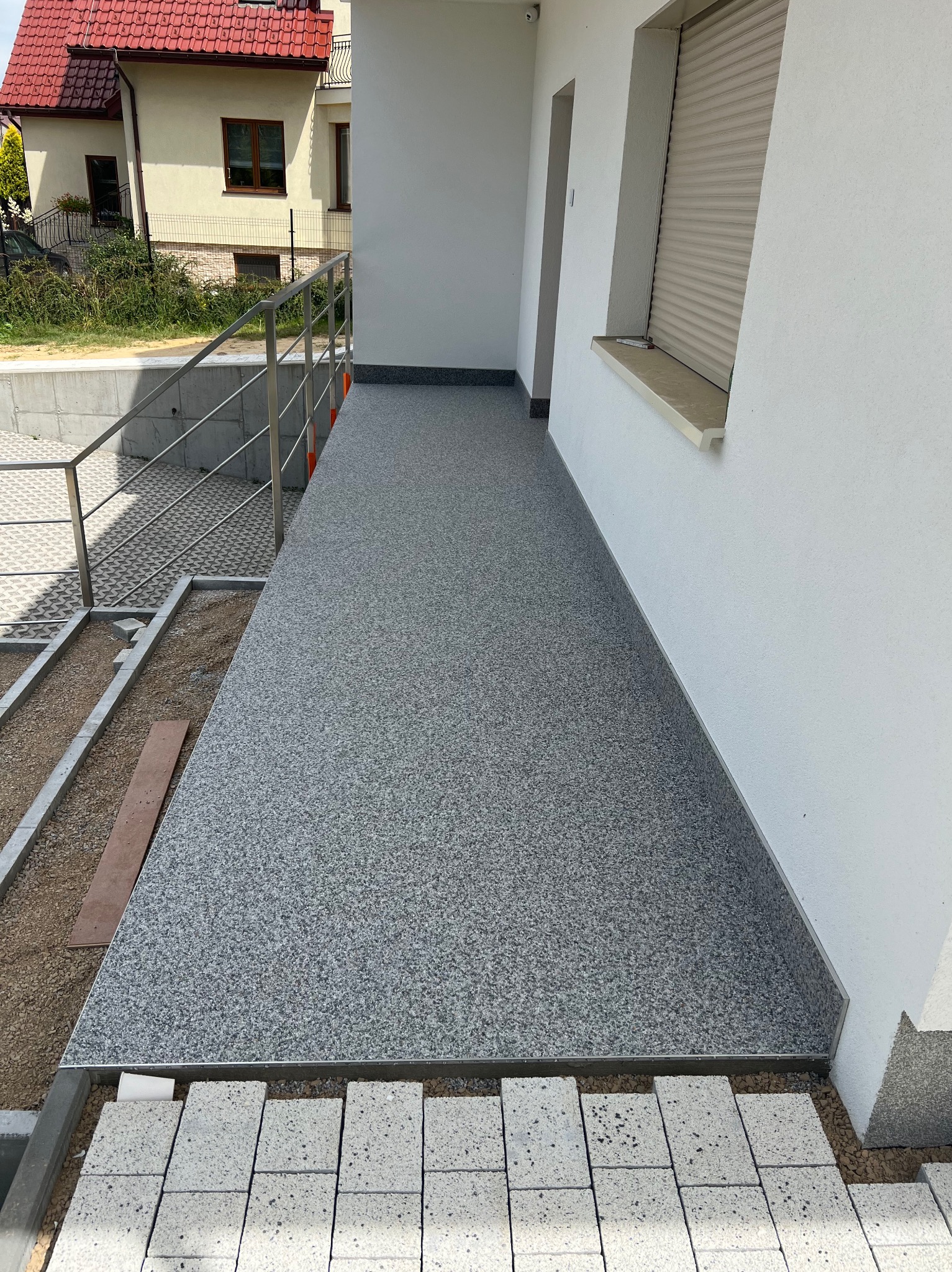 Szara, żywiczna posadzka na zewnątrz budynku, z widocznymi fragmentami przygotowywanej podbudowy z kostki brukowej i metalową balustradą w tle.