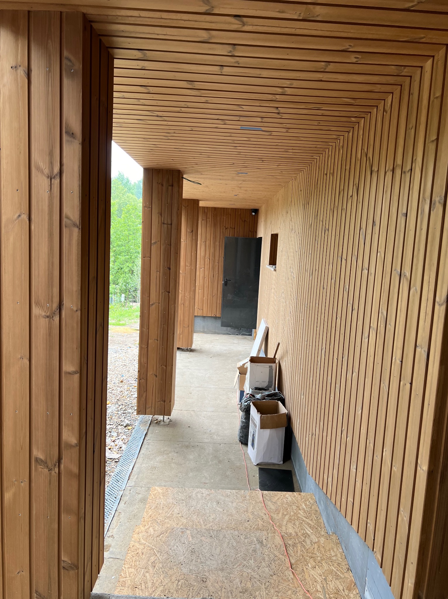 Wnętrze budynku w trakcie wykańczania z boazerią z jasnego drewna na ścianach i suficie, widoczne kartony i materiały budowlane wzdłuż ściany, czarne drzwi w głębi.