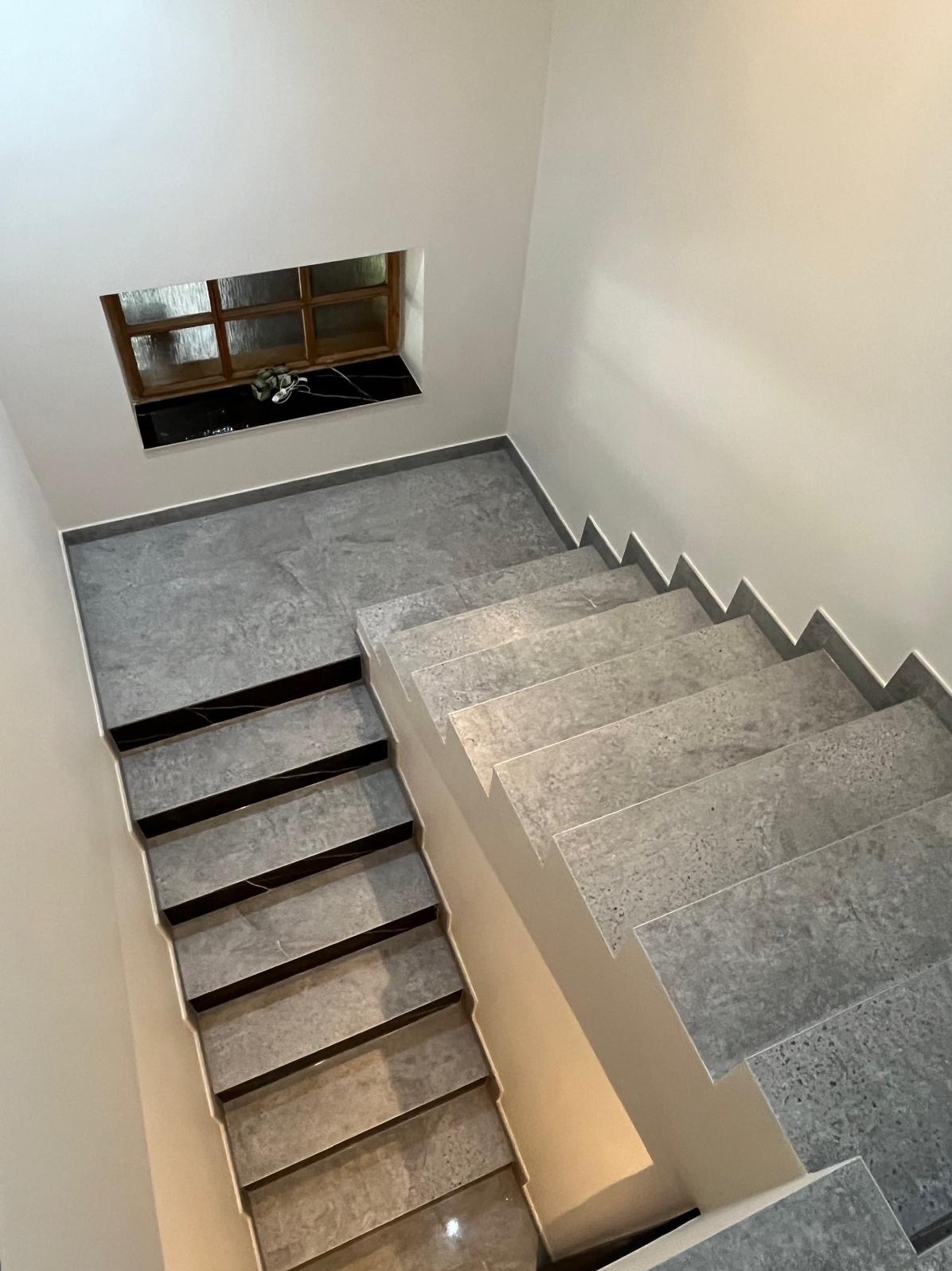 Schody wewnętrzne wykończone szarymi płytkami imitującymi kamień, z kontrastującymi czarnymi wstawkami na stopniach, oświetlone od dołu, z małym okienkiem w tle.