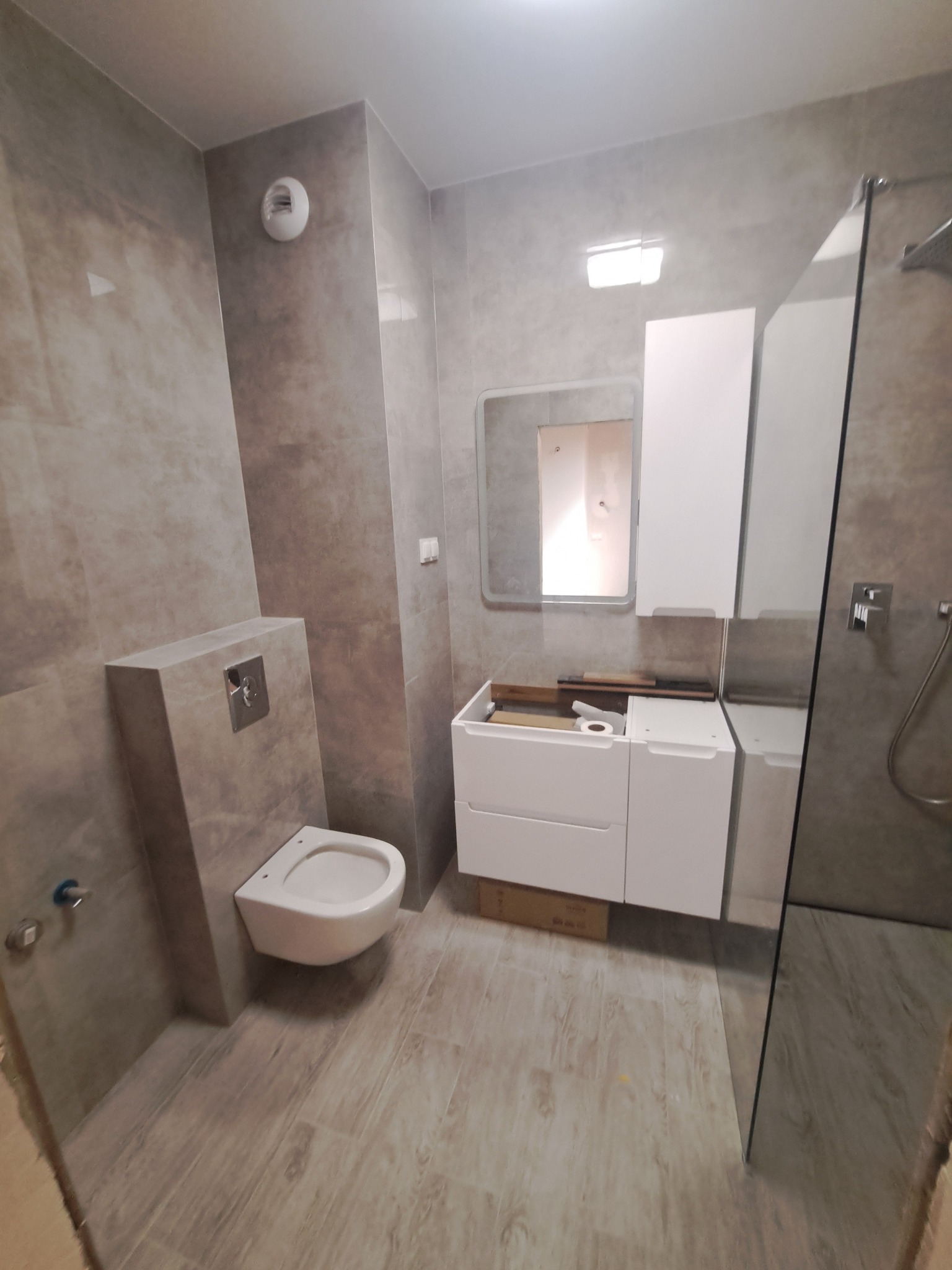 Szara łazienka w trakcie wykańczania, z podwieszaną toaletą, umywalką z szafką i kabiną prysznicową ze szklaną ścianką, widoczne elementy montażowe i karton pod szafką.