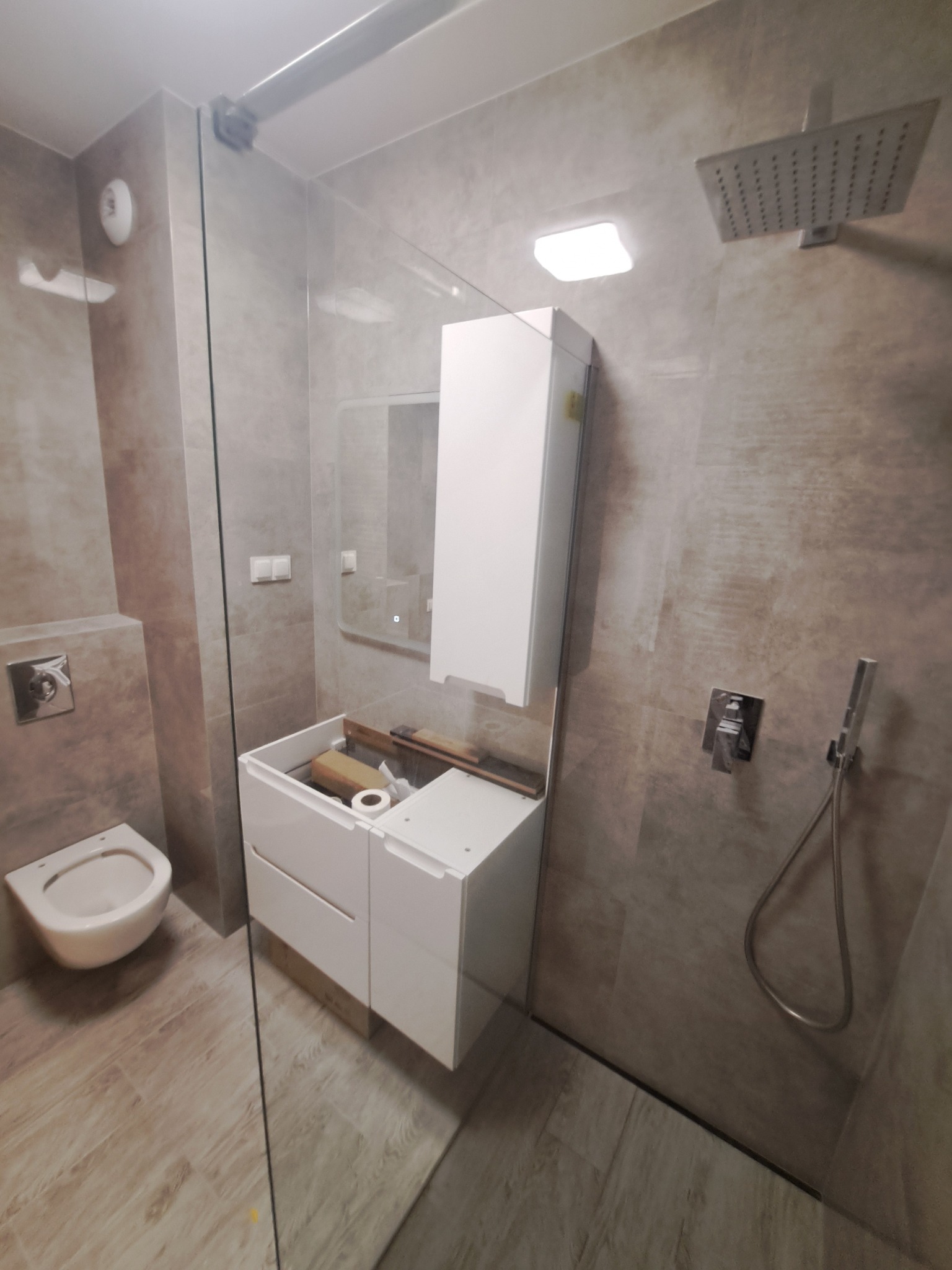 Montaż szafki pod umywalkę w łazience z kabiną prysznicową typu walk-in, widoczne narzędzia i rolka papieru toaletowego wewnątrz szafki, płytki imitujące drewno na podłodze.