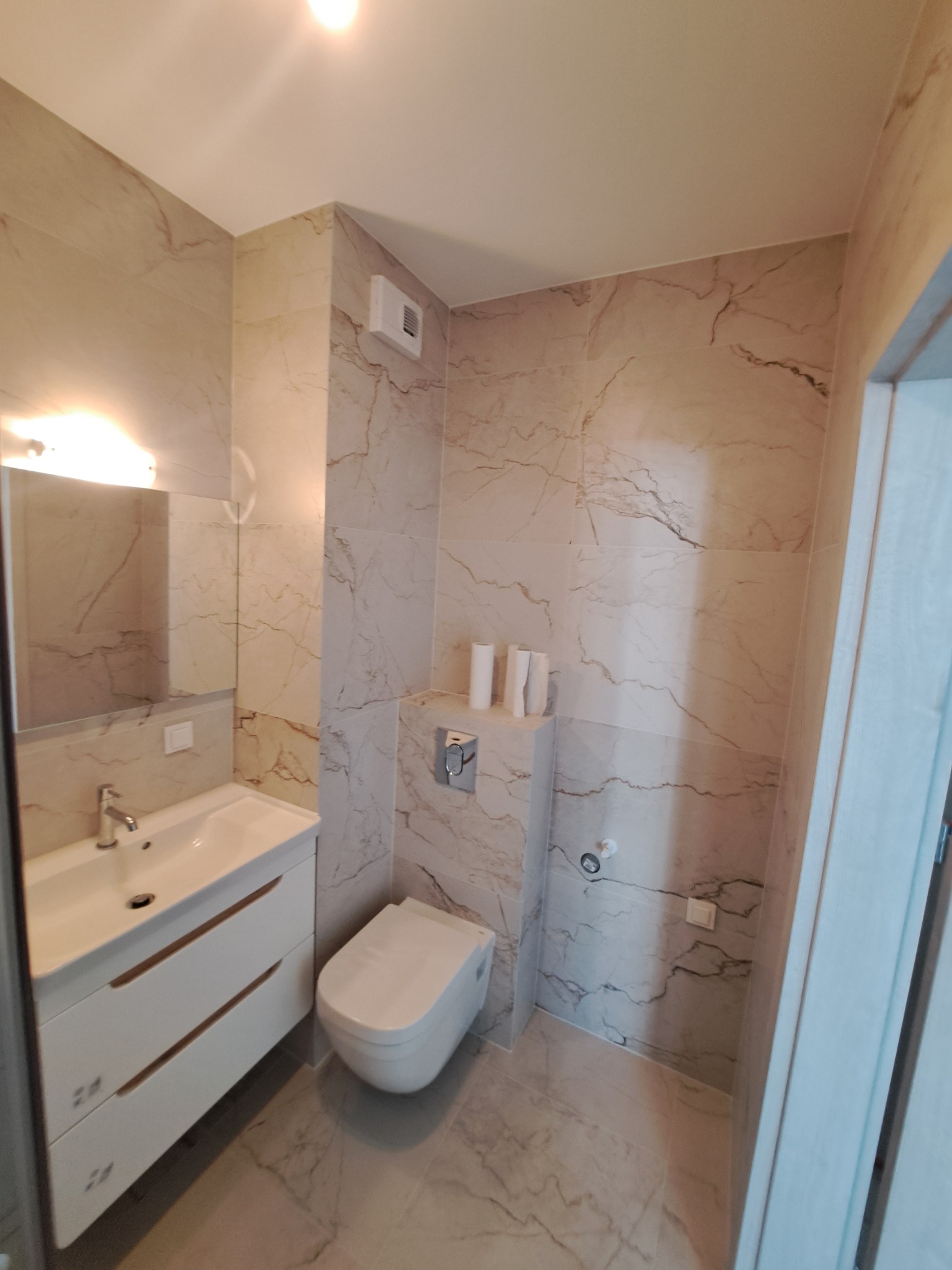 Wykończona łazienka w jasnych marmurkowych płytkach z wiszącą toaletą, umywalką z szafką i lustrem.