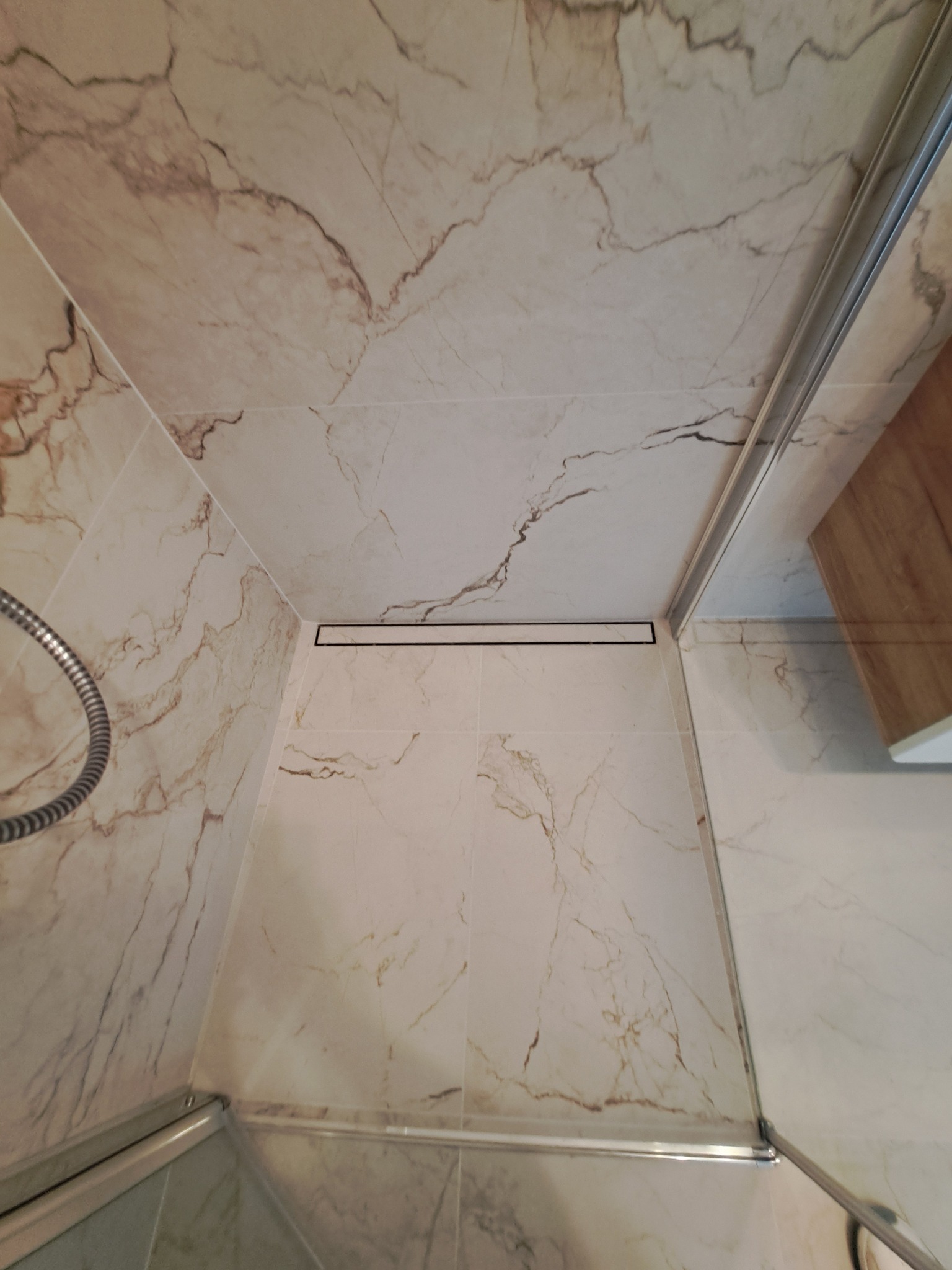 Kabina prysznicowa wykończona płytkami imitującymi marmur, z odpływem liniowym w podłodze i chromowaną słuchawką prysznicową.