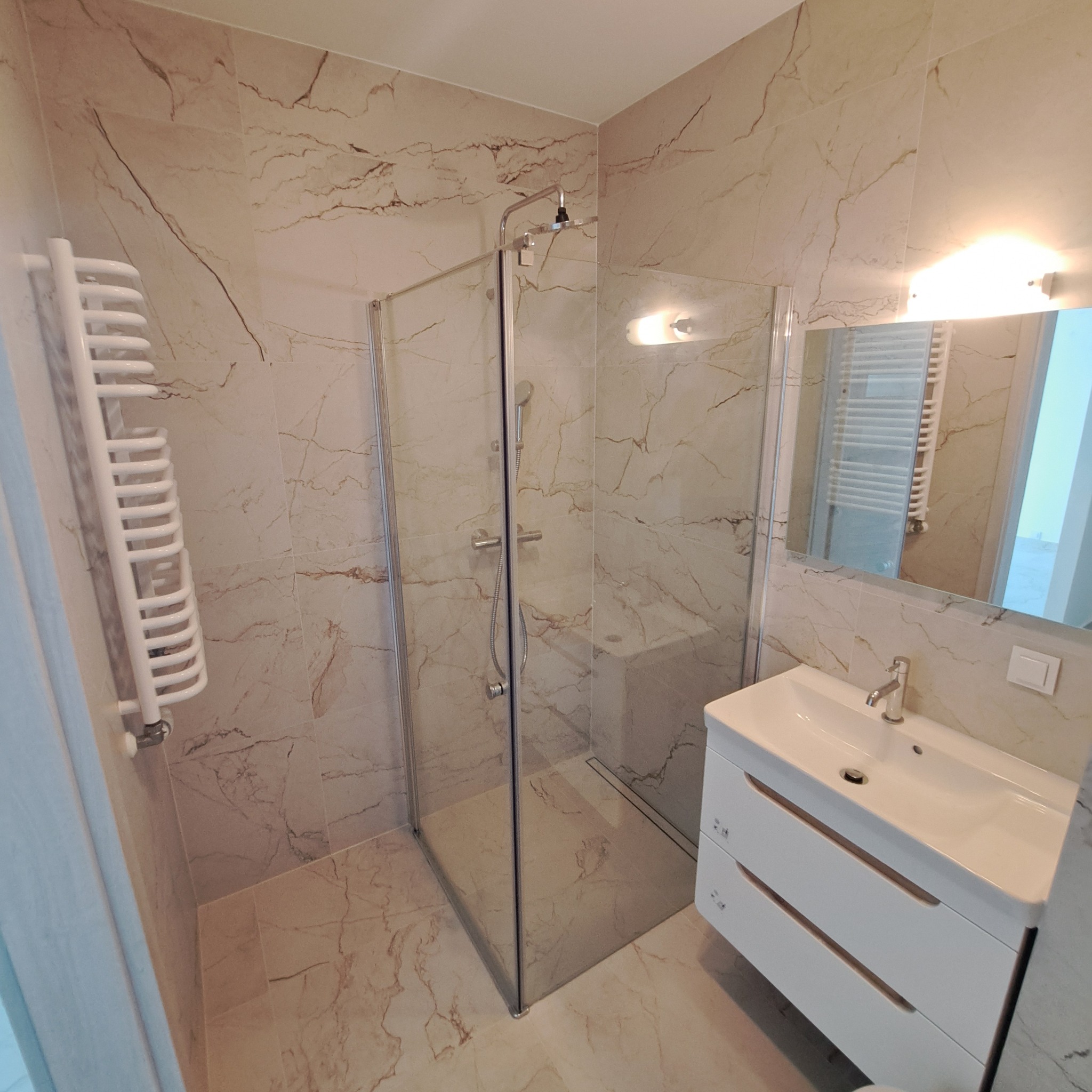 Nowoczesna łazienka z kabiną prysznicową typu walk-in, umywalką z szafką z dwiema szufladami oraz grzejnikiem drabinkowym, wykończona płytkami imitującymi marmur.
