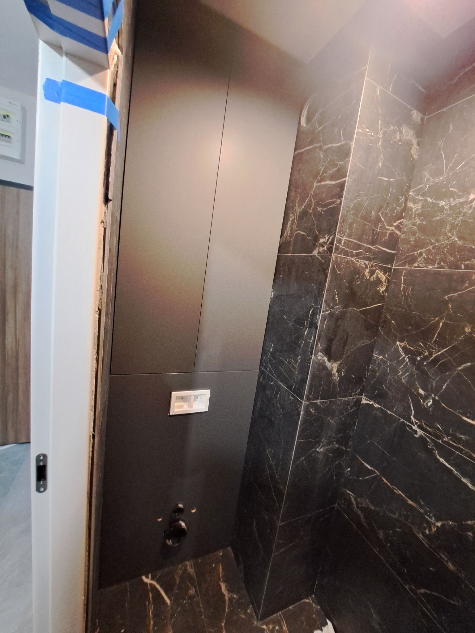 Częściowo wykończona łazienka z czarnymi płytkami imitującymi marmur, widoczny fragment instalacji sanitarnej i rama drzwi zabezpieczona niebieską taśmą malarską.