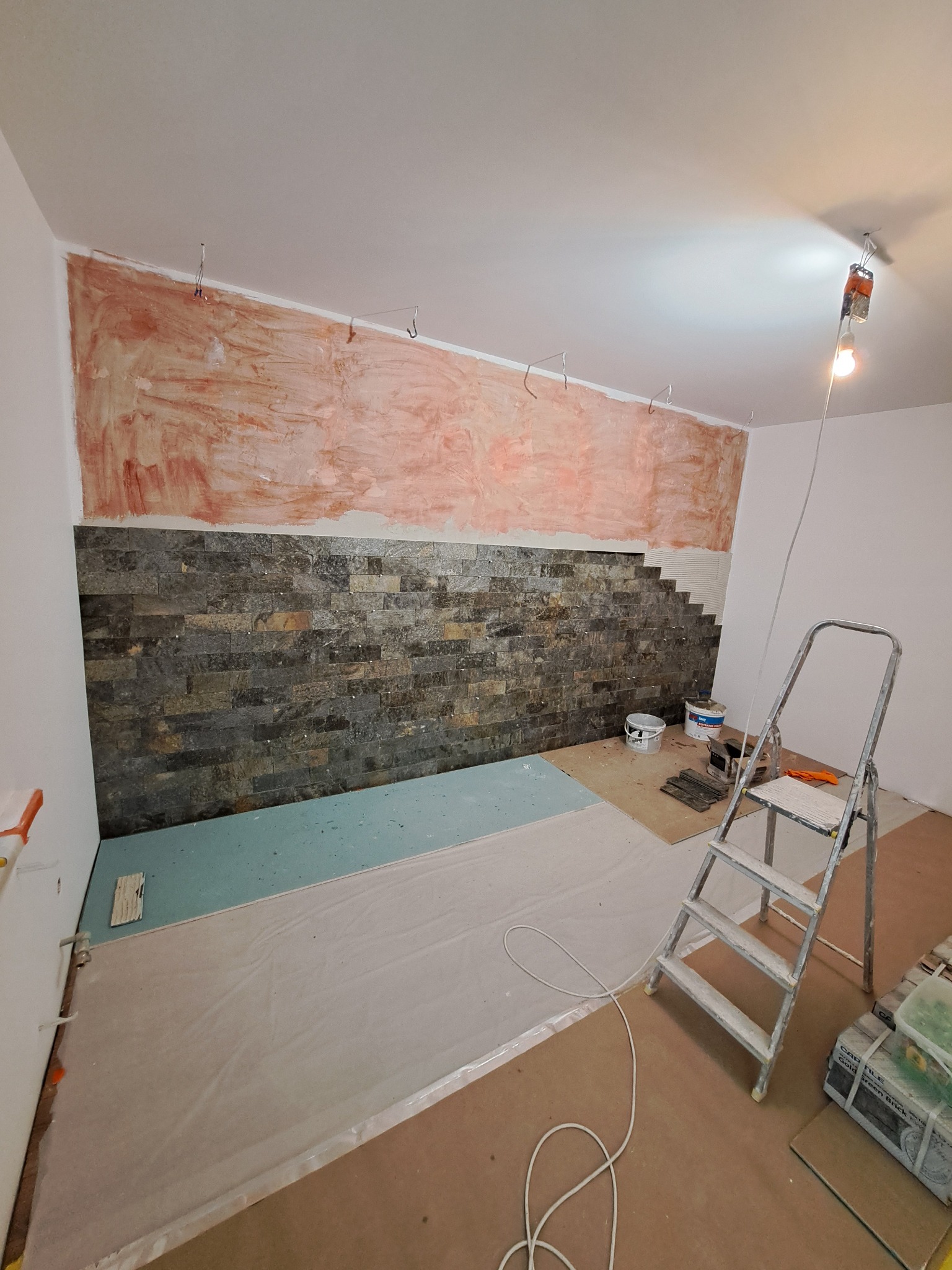 Ściana w trakcie wykańczania z częściowo położoną okładziną z kamienia dekoracyjnego, widoczne ślady po szpachlowaniu na górze ściany, drabina, wiaderka z farbami i narzędzia na podłodze...