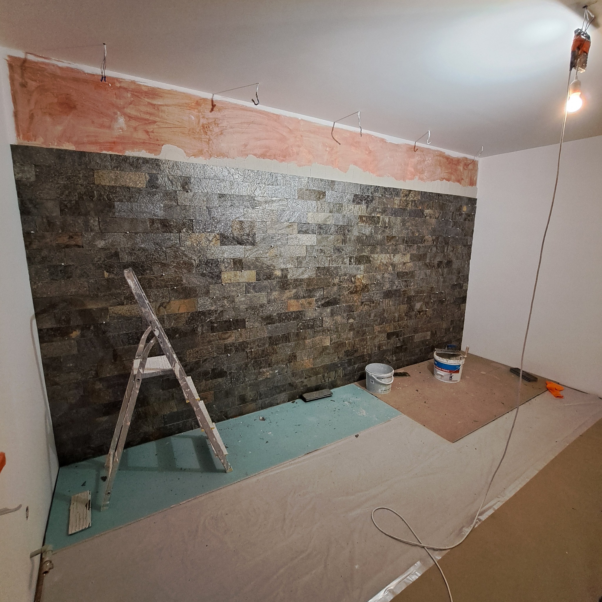 Ściana z ciemnej, kamiennej okładziny w trakcie montażu, widoczne ślady po szpachlowaniu na górze ściany, drabina, wiaderka z klejem i zabezpieczona podłoga.