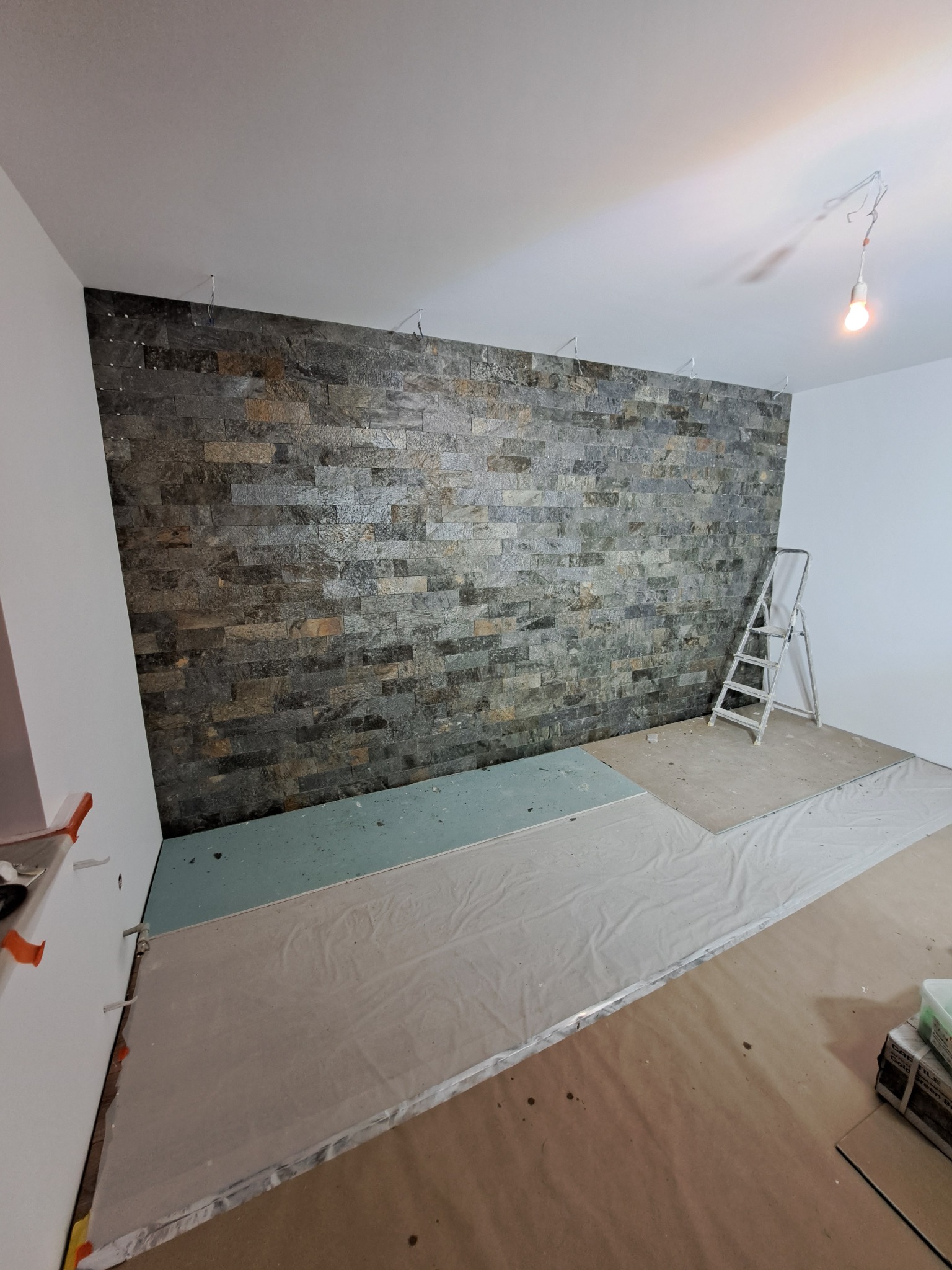Ściana wyłożona dekoracyjnym kamieniem w odcieniach szarości i brązu, z widocznymi przewodami elektrycznymi u góry, przygotowana do dalszych prac wykończeniowych, drabina oparta o ścianę, podłoga...