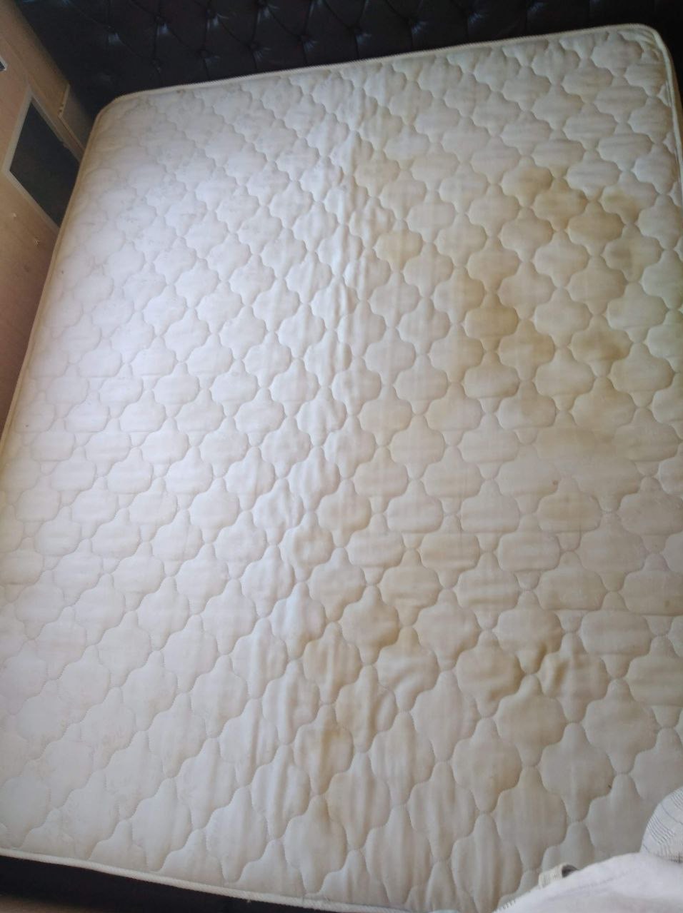 Brudny materac z widocznymi plamami na jasnym, pikowanym poszyciu, leżący na łóżku z ciemnym zagłówkiem.