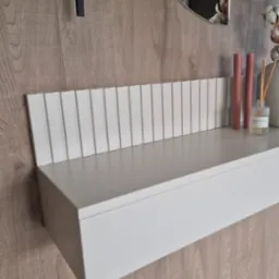 Biała, wisząca konsola z ryflowanym panelem, dekorowana świecami i dyfuzorem zapachowym, na tle drewnianej ściany z okrągłym lustrem.