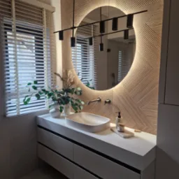 Nowoczesna łazienka z umywalką nablatową, szafką z szufladami w kolorze szarym, okrągłym lustrem z podświetleniem LED i dekoracyjną ścianą z drewnopodobnych paneli 3D.