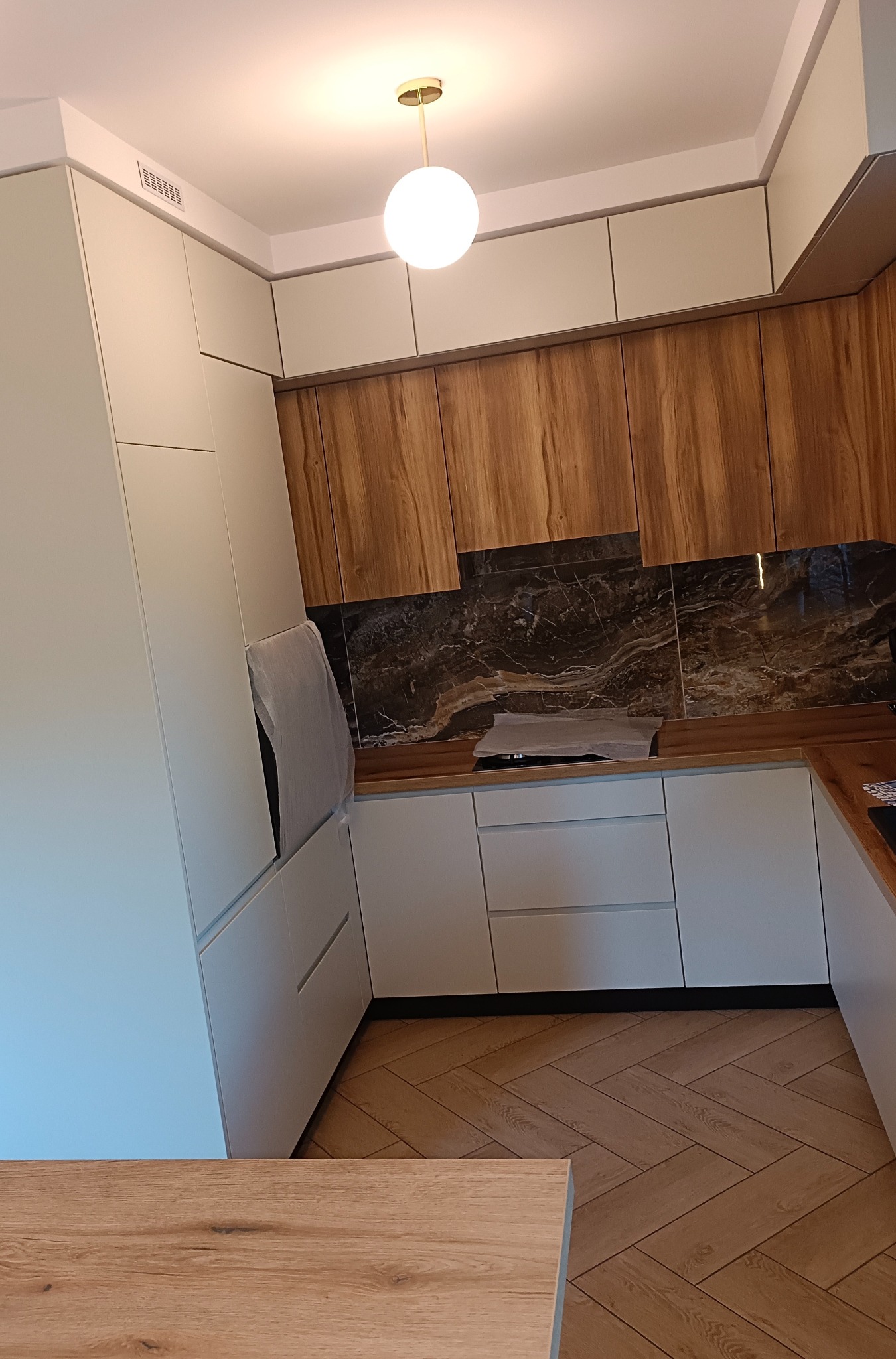 Nowoczesna kuchnia z drewnianymi frontami szafek wiszących i jasnymi, gładkimi frontami szafek dolnych oraz wysokiej zabudowy, parkiet ułożony w jodełkę, okrągła lampa sufitowa.