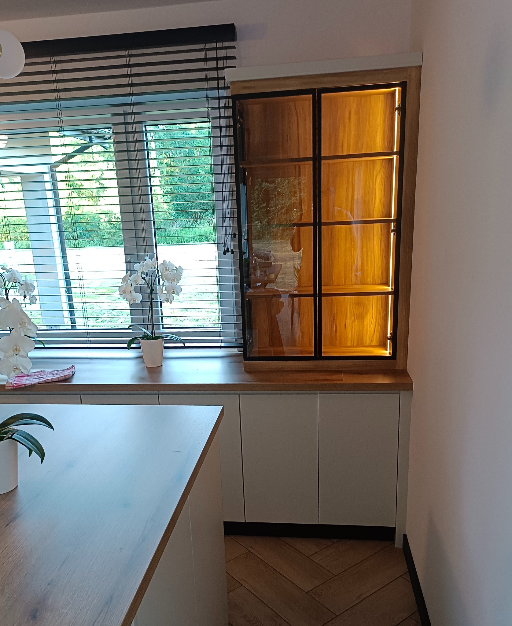 Wbudowana przeszklona witryna z oświetleniem LED, umieszczona nad szafkami kuchennymi, z widocznymi żaluzjami okiennymi i roślinami doniczkowymi w tle.