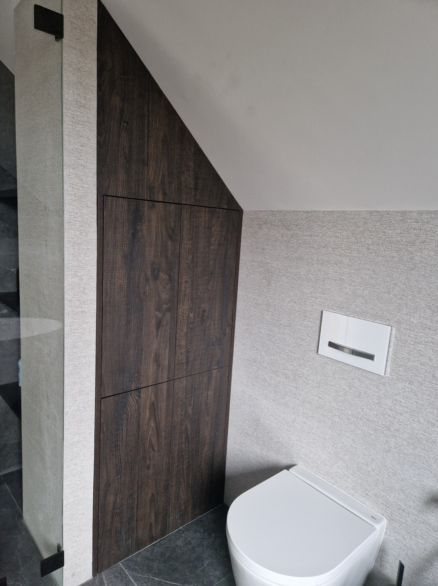 Szafka wnękowa z ciemnego drewna w łazience ze spłuczką podtynkową i toaletą wiszącą.