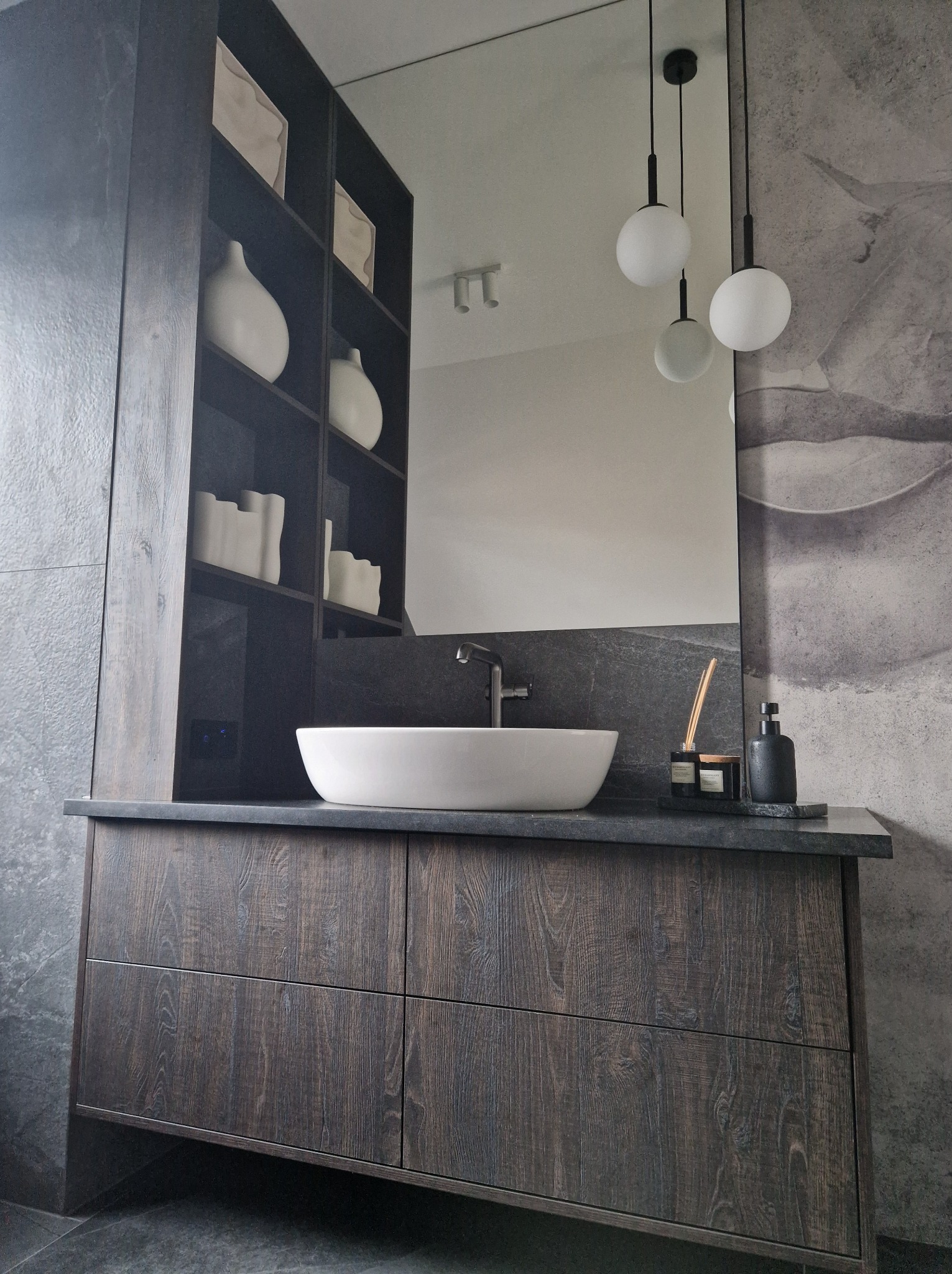 Nowoczesna łazienka z umywalką nablatową, szafką z ciemnego drewna i otwartymi półkami z dekoracyjnymi wazonami, oświetlona trzema kulistymi lampami wiszącymi.