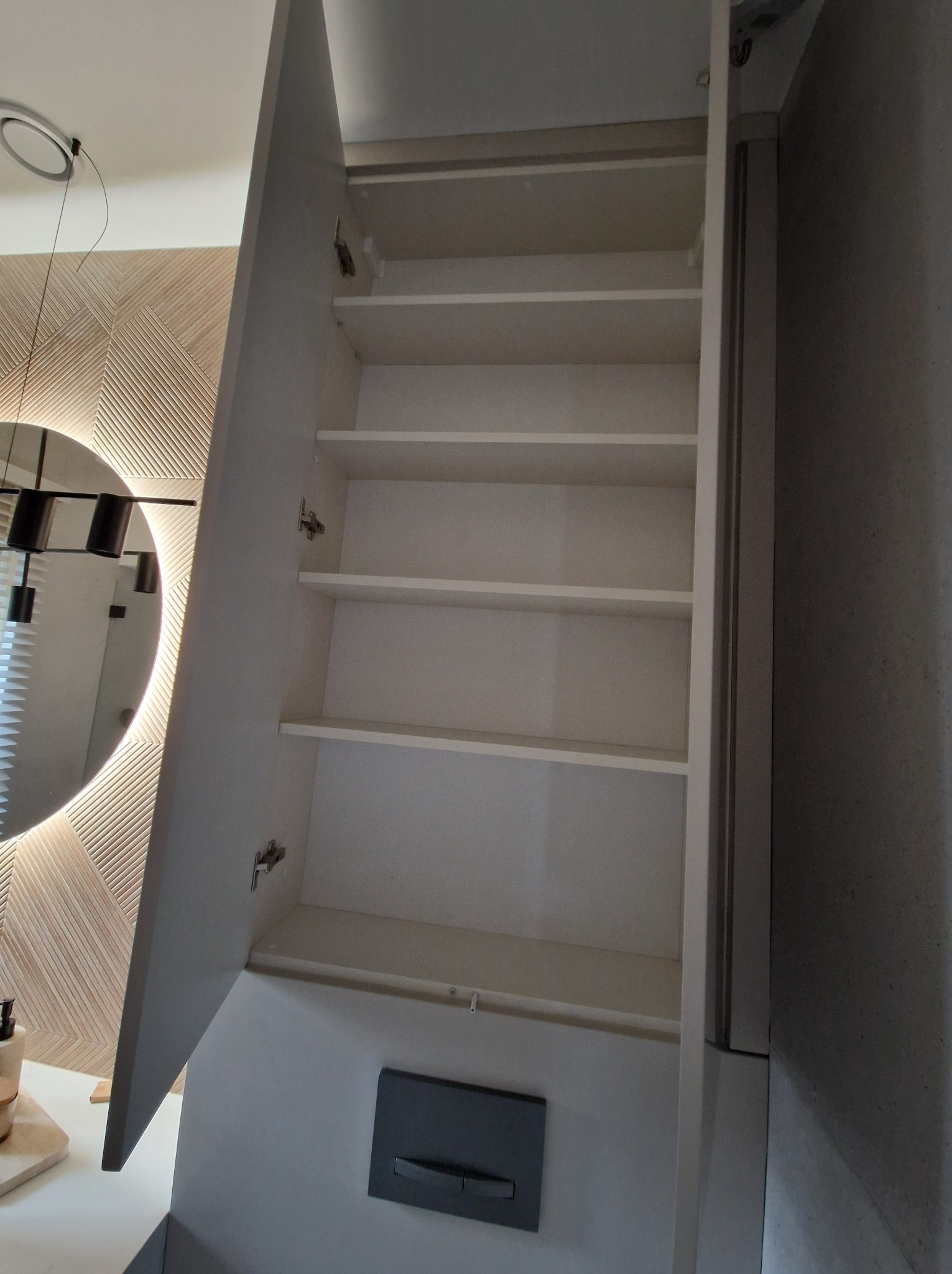 Otwarta szafka łazienkowa z półkami, widoczne zawiasy, na ścianie dekoracyjny panel z drewna i okrągłe lustro z podświetleniem LED.