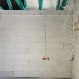 Surowa ściana z betonu komórkowego z rozprowadzoną instalacją elektryczną: kable w peszlach i puszki pod gniazdka. Widoczne elementy konstrukcji dachu.