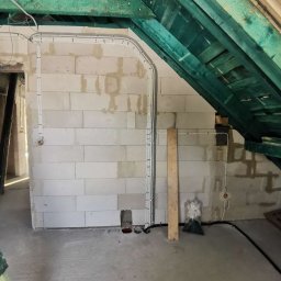 Pa tryk - Ściana z pustaków z poprowadzonymi przewodami elektrycznymi w szarych rurkach. Zielona drewniana konstrukcja dachu. W tle otwór drzwiowy i fragment drabiny.