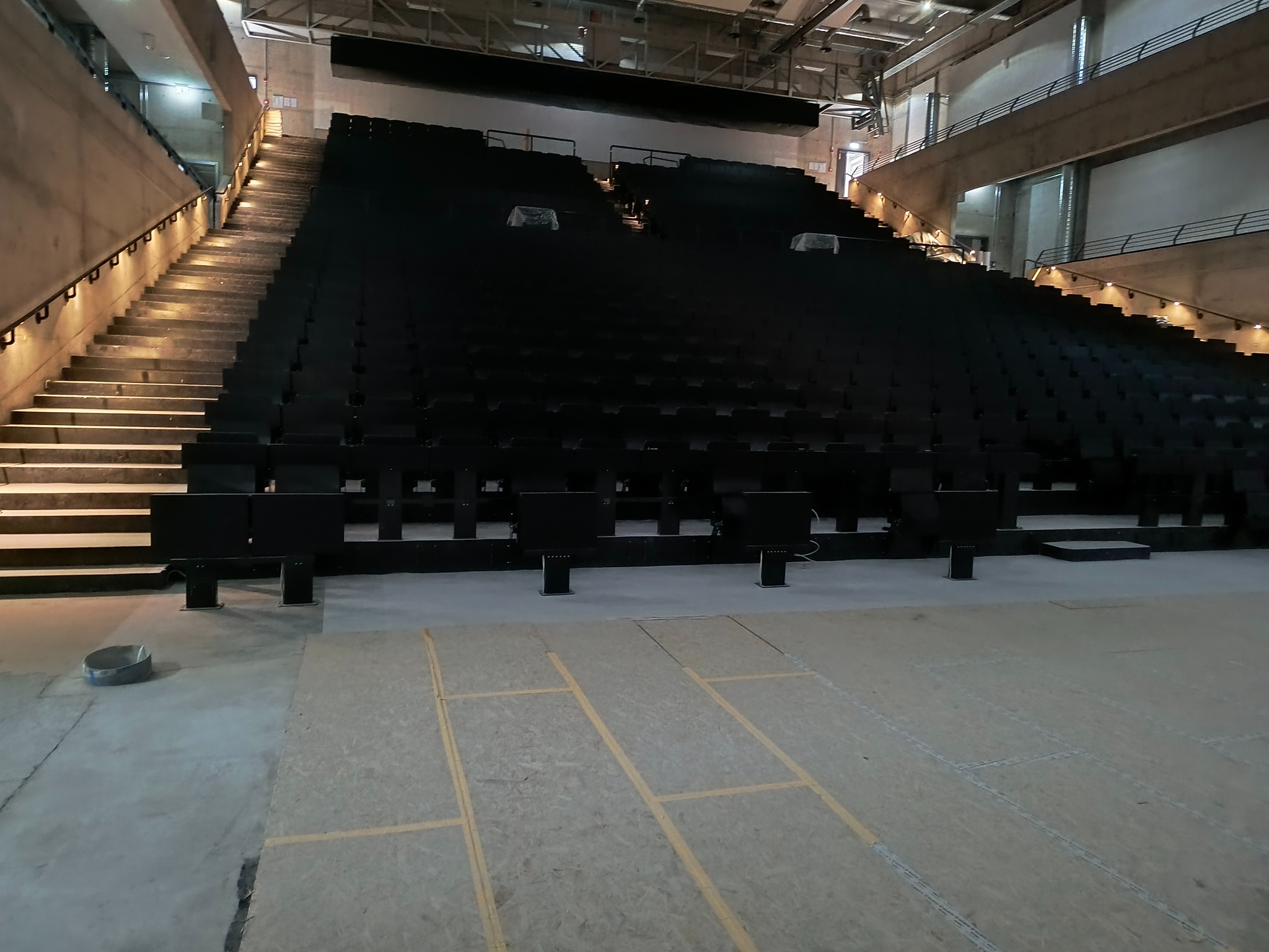 Sala widowiskowa z rzędami czarnych foteli i betonowymi schodami z oświetleniem. Na podłodze widoczne linie wyznaczające układ. W tle surowy, betonowy wystrój.