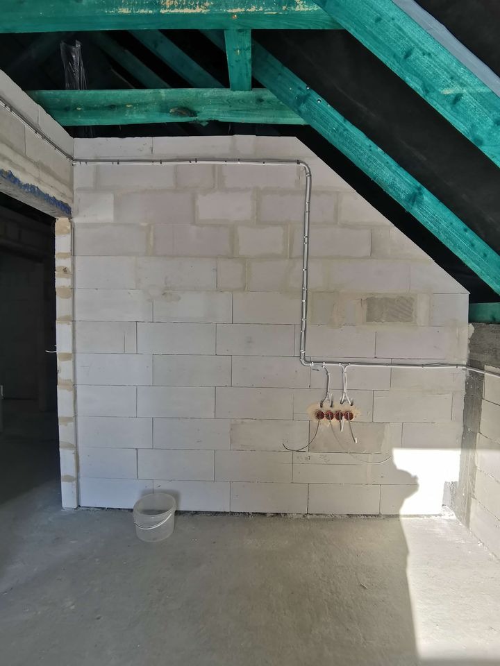 Surowa ściana z gazobetonu z poprowadzoną, zewnętrzną instalacją elektryczną pod stropem z zielonymi belkami. Widoczne kable w peszlach i puszki pod gniazdka.