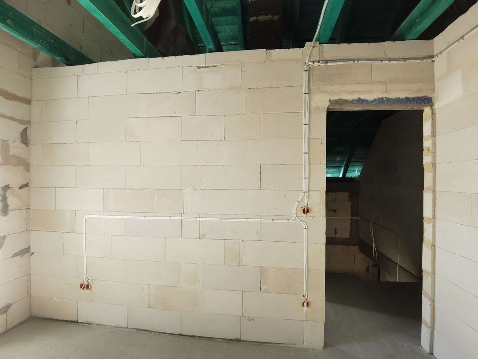 Surowy stan: białe ściany z betonu komórkowego, instalacja elektryczna w peszlu i puszki podtynkowe na ścianie. Widoczne zielone belki stropowe.