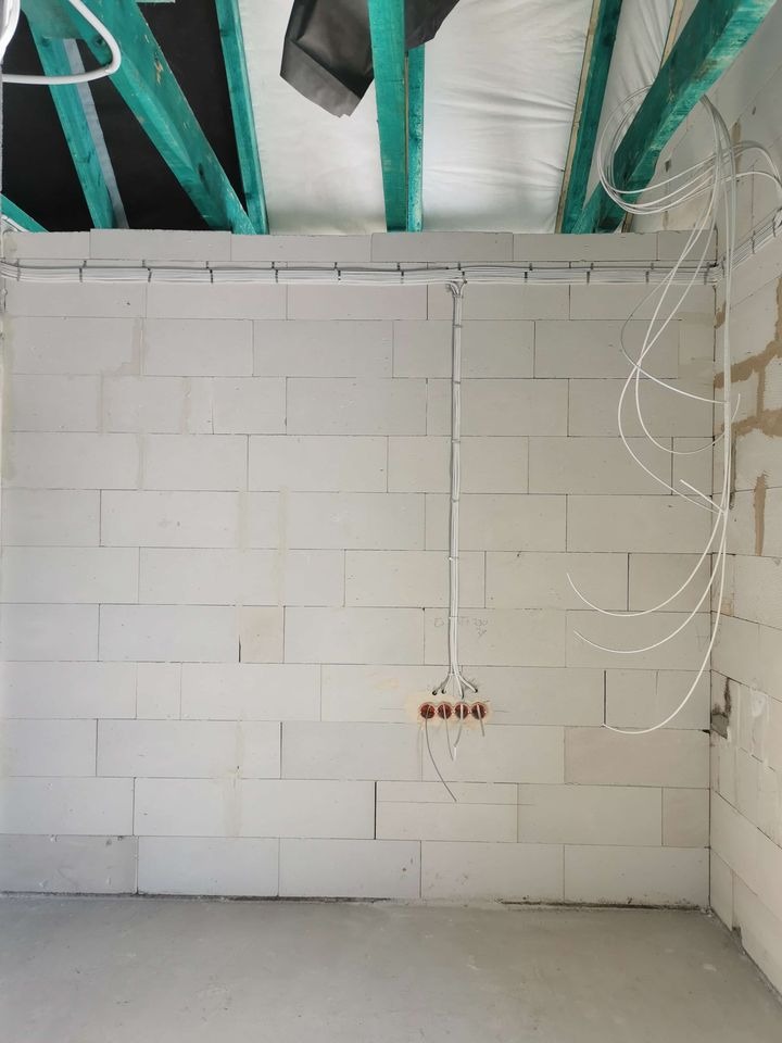 Surowa ściana z betonu komórkowego z rozprowadzoną instalacją elektryczną: kable w peszlach i puszki pod gniazdka. Widoczne elementy konstrukcji dachu.