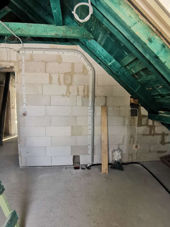 Ściana z pustaków z poprowadzonymi przewodami elektrycznymi w szarych rurkach. Zielona drewniana konstrukcja dachu. W tle otwór drzwiowy i fragment drabiny.