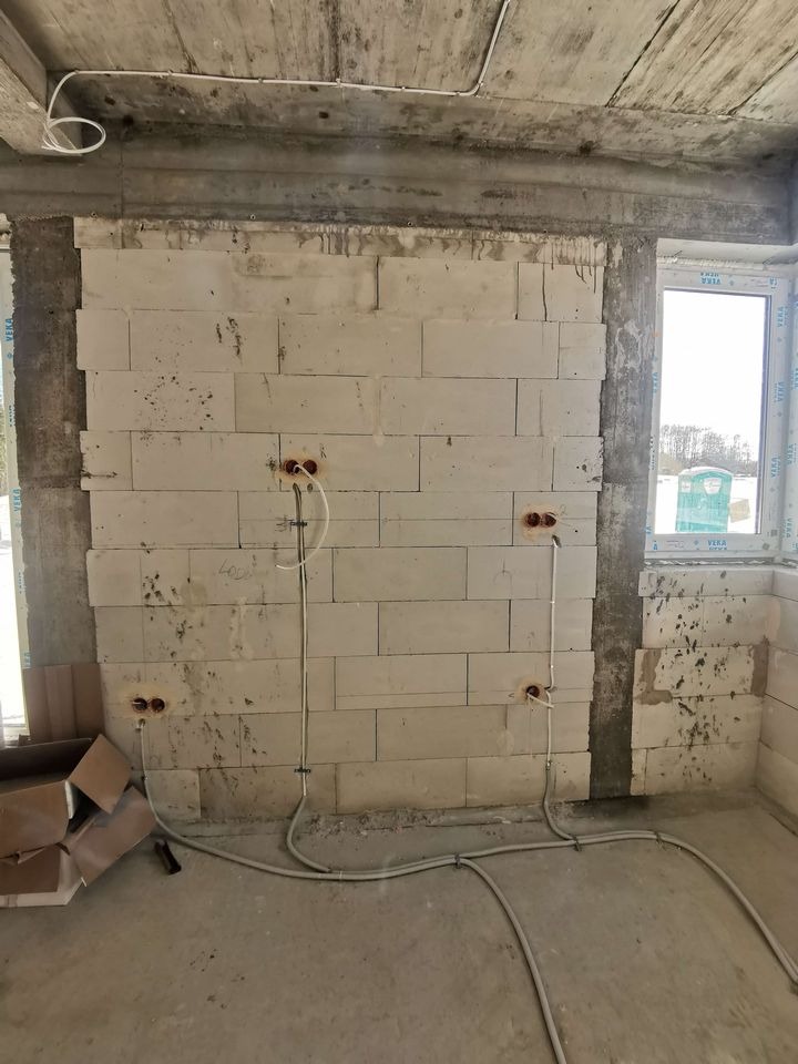 Surowy mur z instalacją elektryczną: przewody w peszlach prowadzone wzdłuż ściany, puszki elektryczne. Widok wnętrza w stanie deweloperskim, okno w tle.