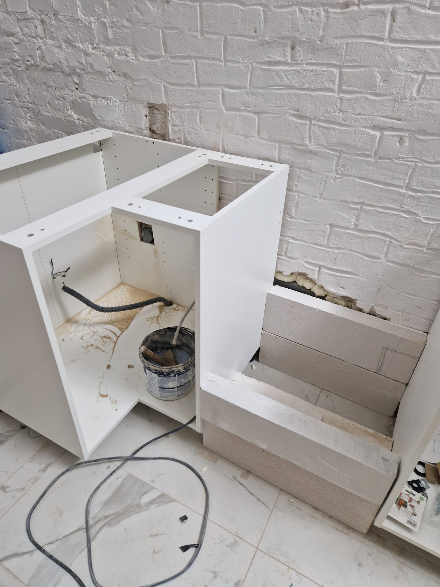 Montaż szafki kuchennej: widoczne białe korpusy szafek, wiadro z zaprawą malarską, przewód elektryczny oraz fragment ściany z surowej cegły i fragmentem zamurowanego otworu.