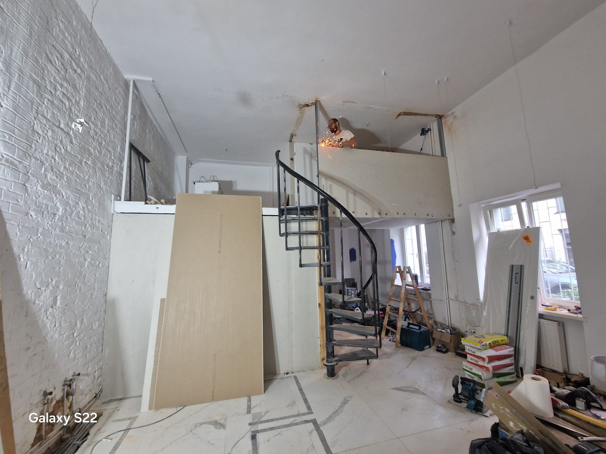 Wnętrze w trakcie remontu z widoczną spiralną klatką schodową, płytami gipsowo-kartonowymi i pracownikiem spawającym elementy konstrukcyjne na antresoli, iskry podczas spawania.