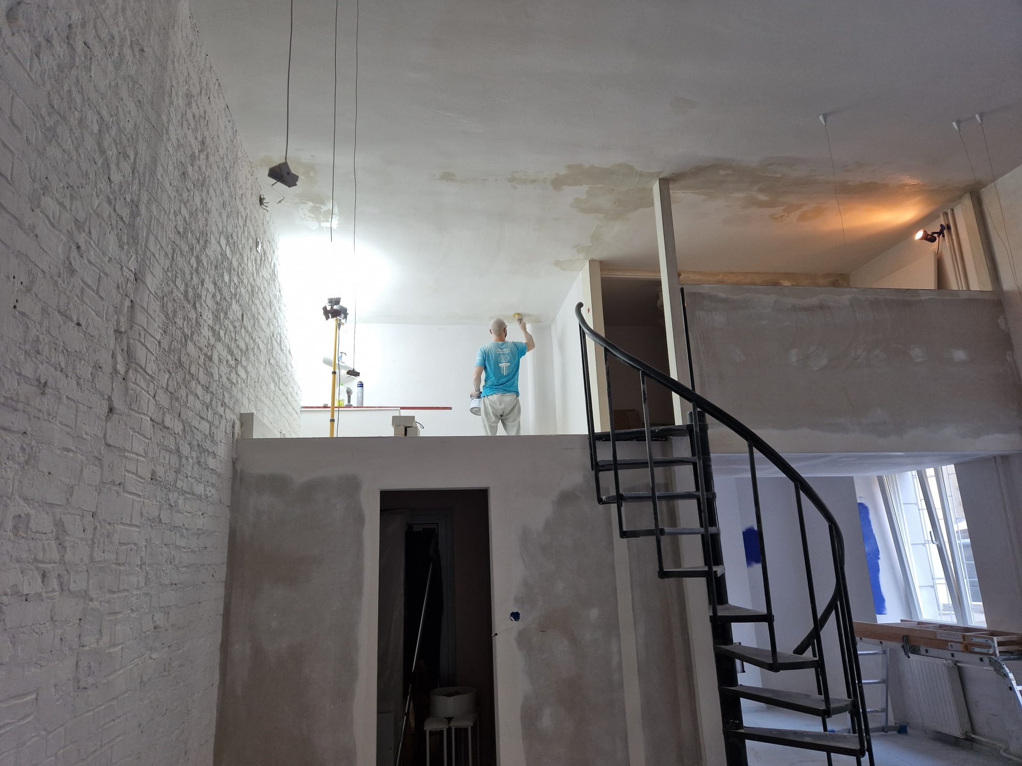 Malowanie sufitu w wysokim pomieszczeniu z antresolą i spiralnymi schodami; widoczny malarz z wałkiem na podwyższeniu, surowa ściana z cegły i ślady po zalaniach na suficie
